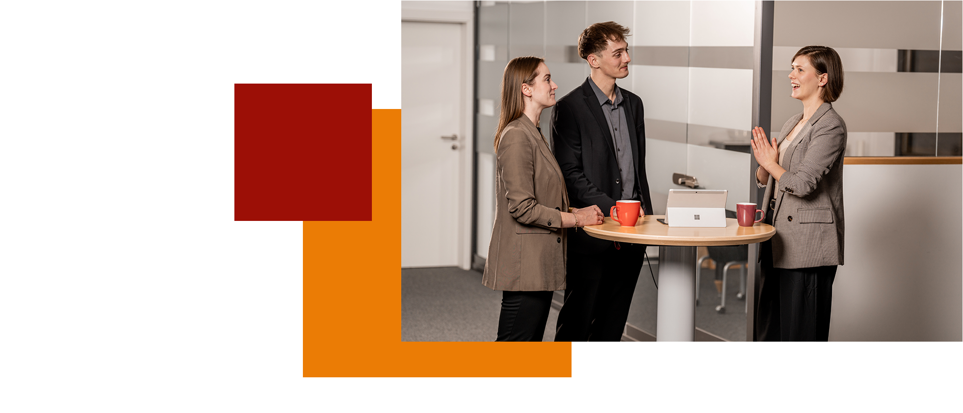 Drei Personen in Business-Kleidung stehen an einem Stehtisch im Büro, eine präsentiert gestikulierend.