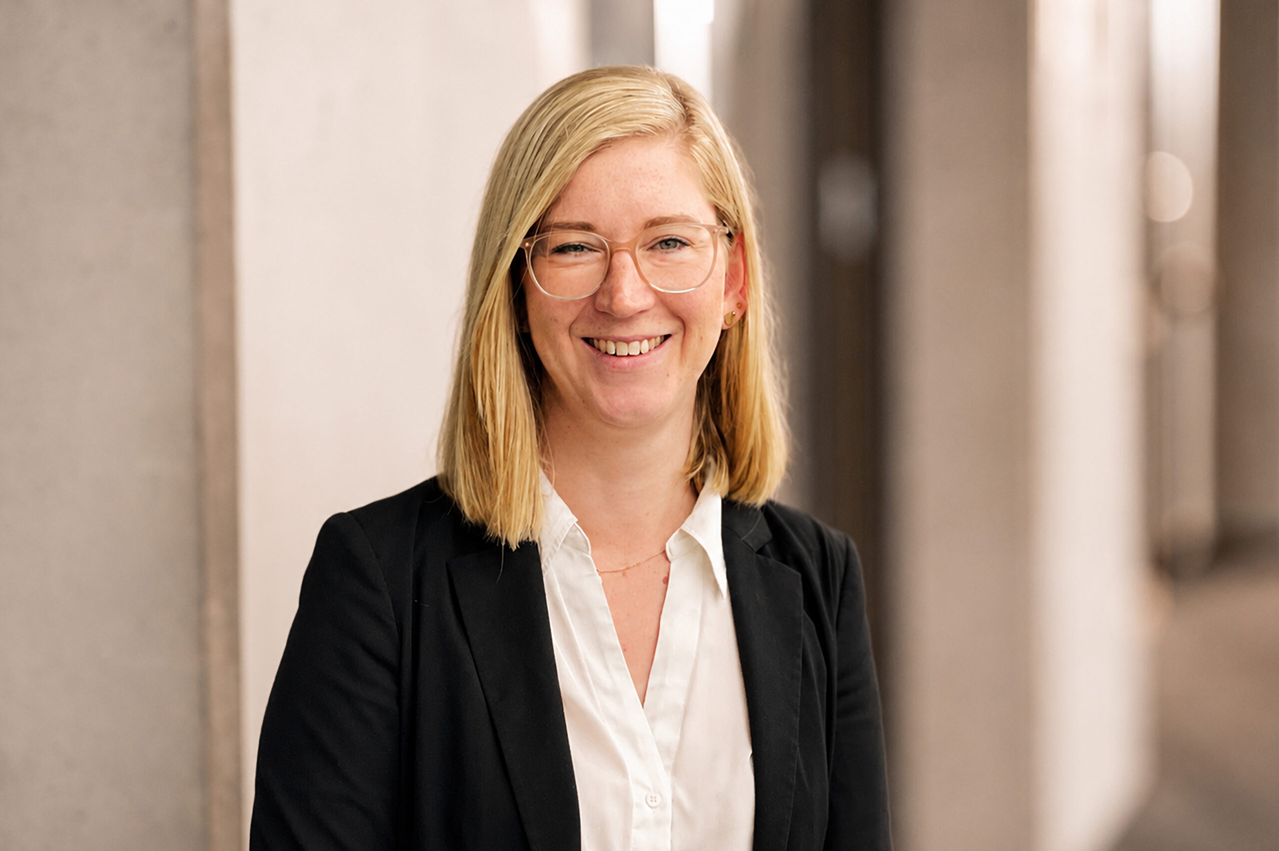Eine lächelnde Frau mit Brille in einem schwarzen Blazer und weißer Bluse in professionellem Umfeld.